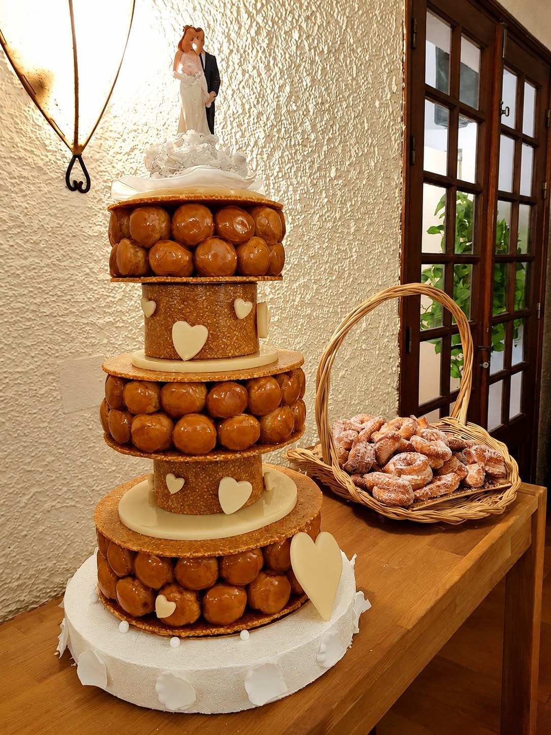 Pièce montée de mariage en choux caramélisés sur trois étages, ornée de cœurs blancs et d'un couple de mariés, avec un panier de beignets sucrés à côté.