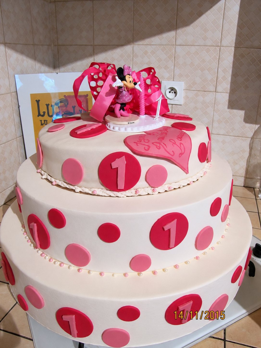 Gâteau d'anniversaire à deux étages décoré de pois roses et rouges, surmonté d'une figurine de Minnie, d'un nœud rose et d'une bougie.