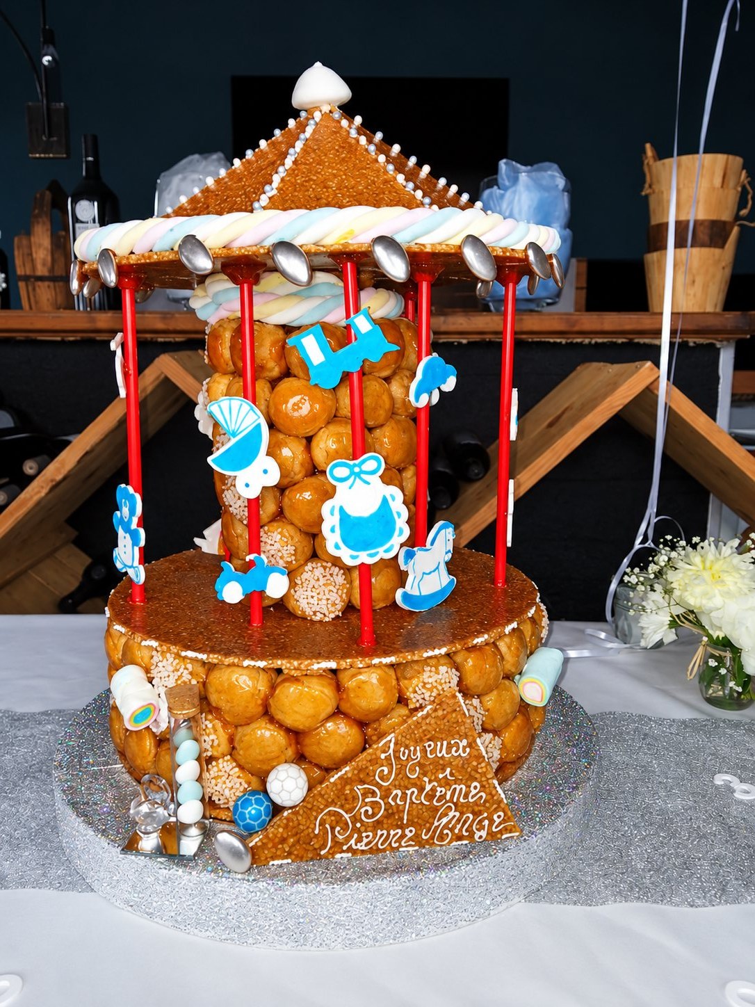Pièce montée de baptême en forme de carrousel, composée de choux caramélisés, de guimauves torsadées et de décorations bleues, posée sur un plateau pailleté.