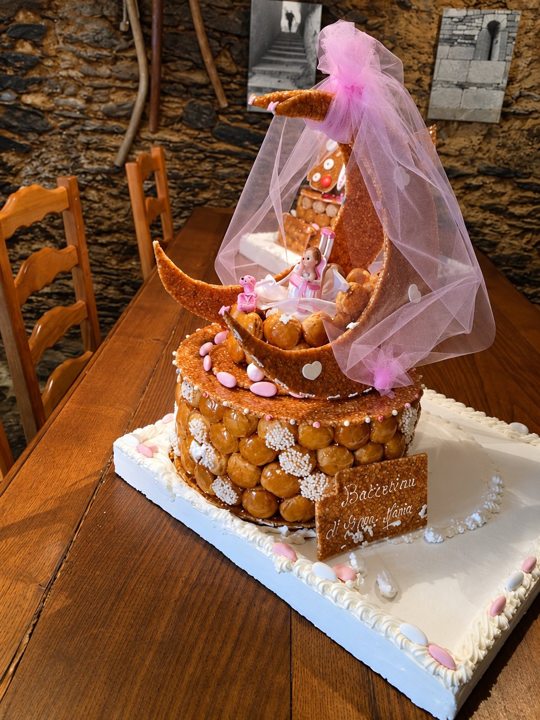 Une pièce montée de choux au caramel, surmontée d'un croissant de lune en pain d'épices et d'un voile rose, est posée sur une table en bois.