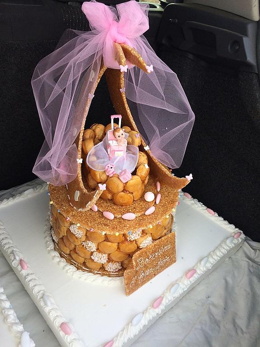 Pièce montée de baptême composée de choux caramélisés, ornée d'un voile de tulle rose, d'un nœud et d'une figurine de bébé, posée sur un plateau dans un coffre de voiture.