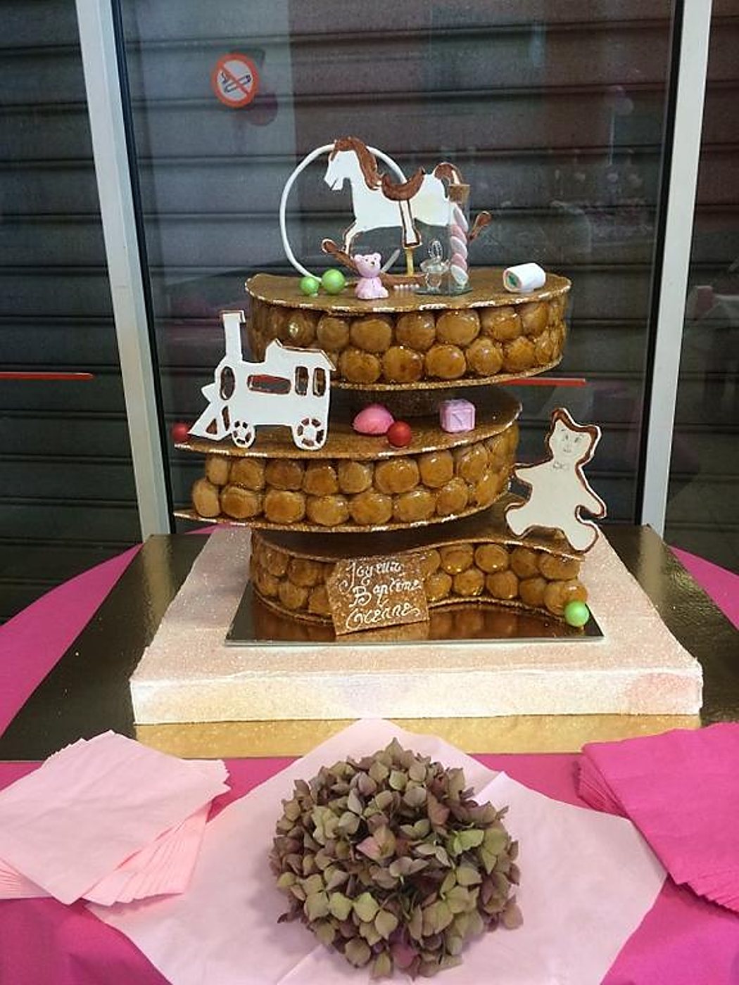 Une pièce montée de choux caramélisés sur trois étages, ornée d'un cheval à bascule, d'un petit train et d'un ourson en chocolat, posée sur une table rose.