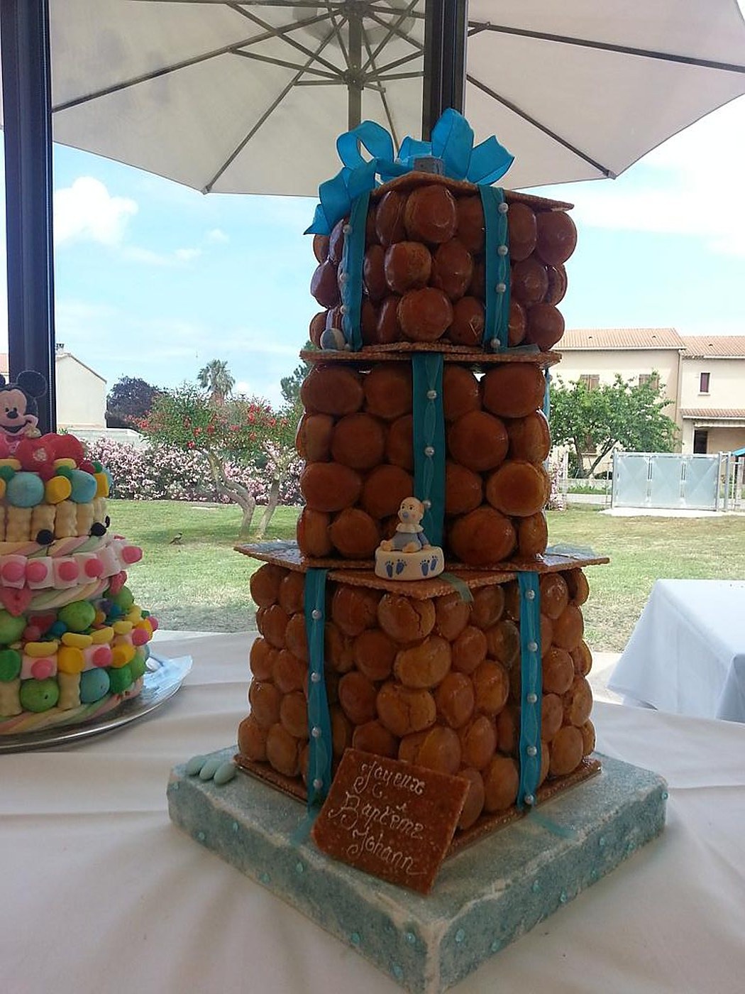 Une pièce montée de choux caramélisés ornée de rubans bleus et d'une plaque de baptême est posée sur une table dans un jardin, près d'un gâteau de bonbons.