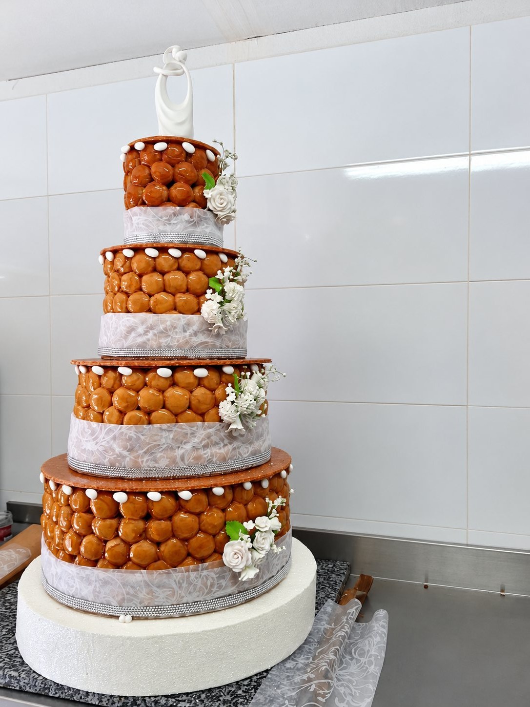 Pièce montée à quatre étages composée de choux caramélisés, ornée de rubans blancs, de strass et de fleurs en sucre blanches, posée sur un plan de travail en granit devant un mur carrelé.