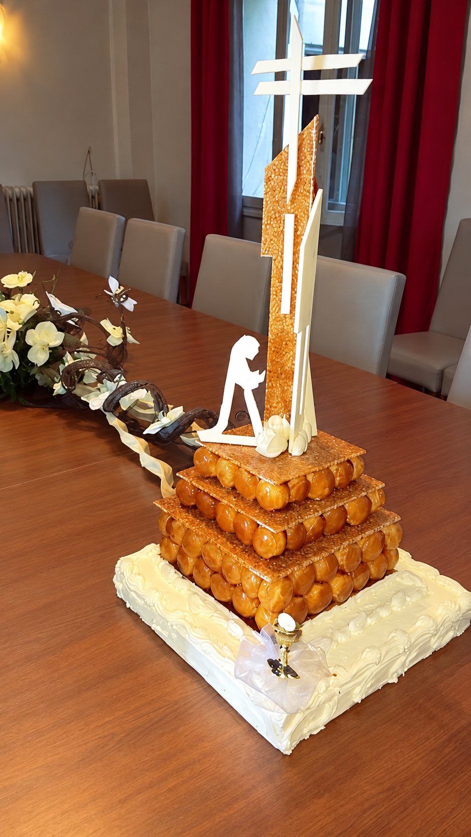 Une pièce montée en étages de choux caramélisés, ornée d'une croix dorée et d'une silhouette blanche, posée sur une table en bois dans une salle à manger.