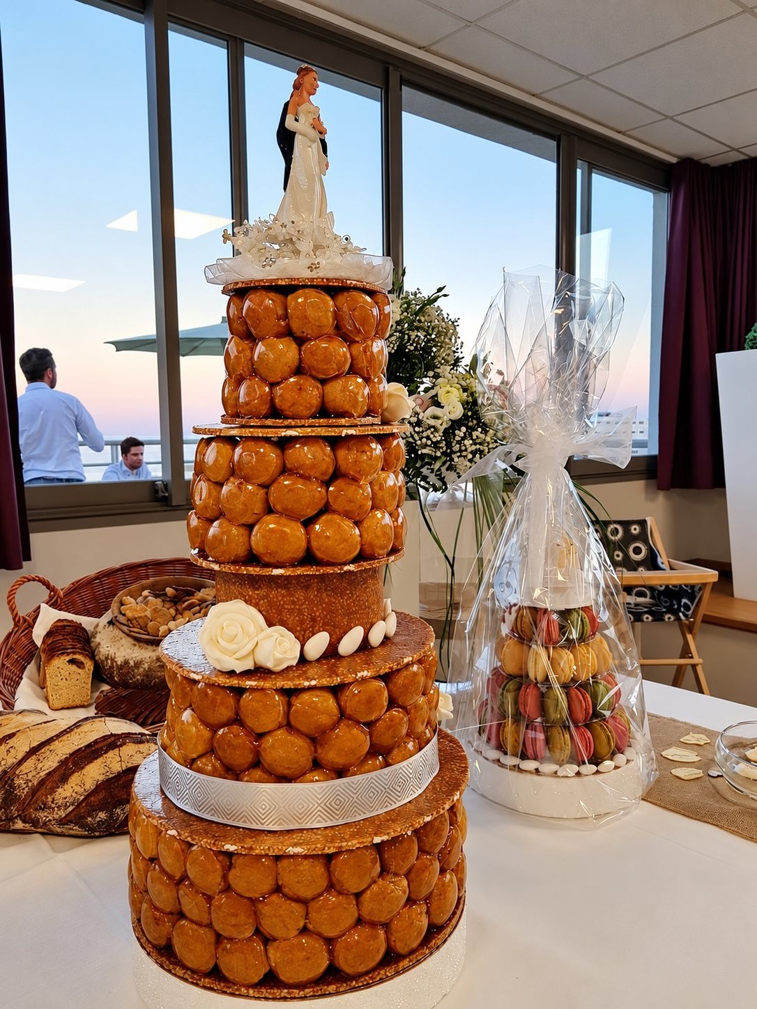 Une pièce montée de choux caramélisés surmontée de figurines de mariés, posée sur une table près d'un bouquet et d'un cylindre de macarons emballé.