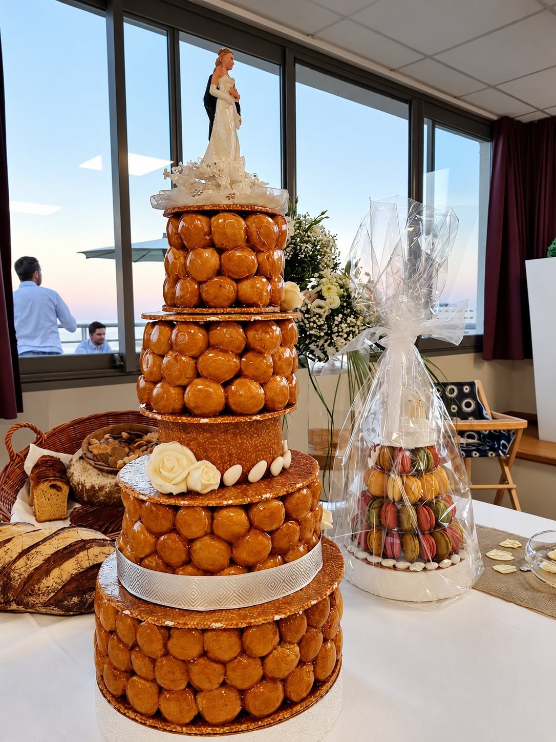 Une pièce montée de mariage composée de choux caramélisés et ornée de roses en pâte d'amandes, avec un présentoir de macarons emballé dans du cellophane et un panier de pain sur une table devant de grandes baies vitrées.