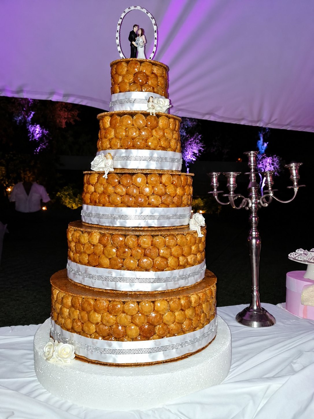 Une pièce montée de mariage à cinq étages, faite de choux caramélisés ornés de rubans blancs et de roses en sucre, surmontée de figurines de mariés et posée sur une table nappée de blanc près d'un chandelier.