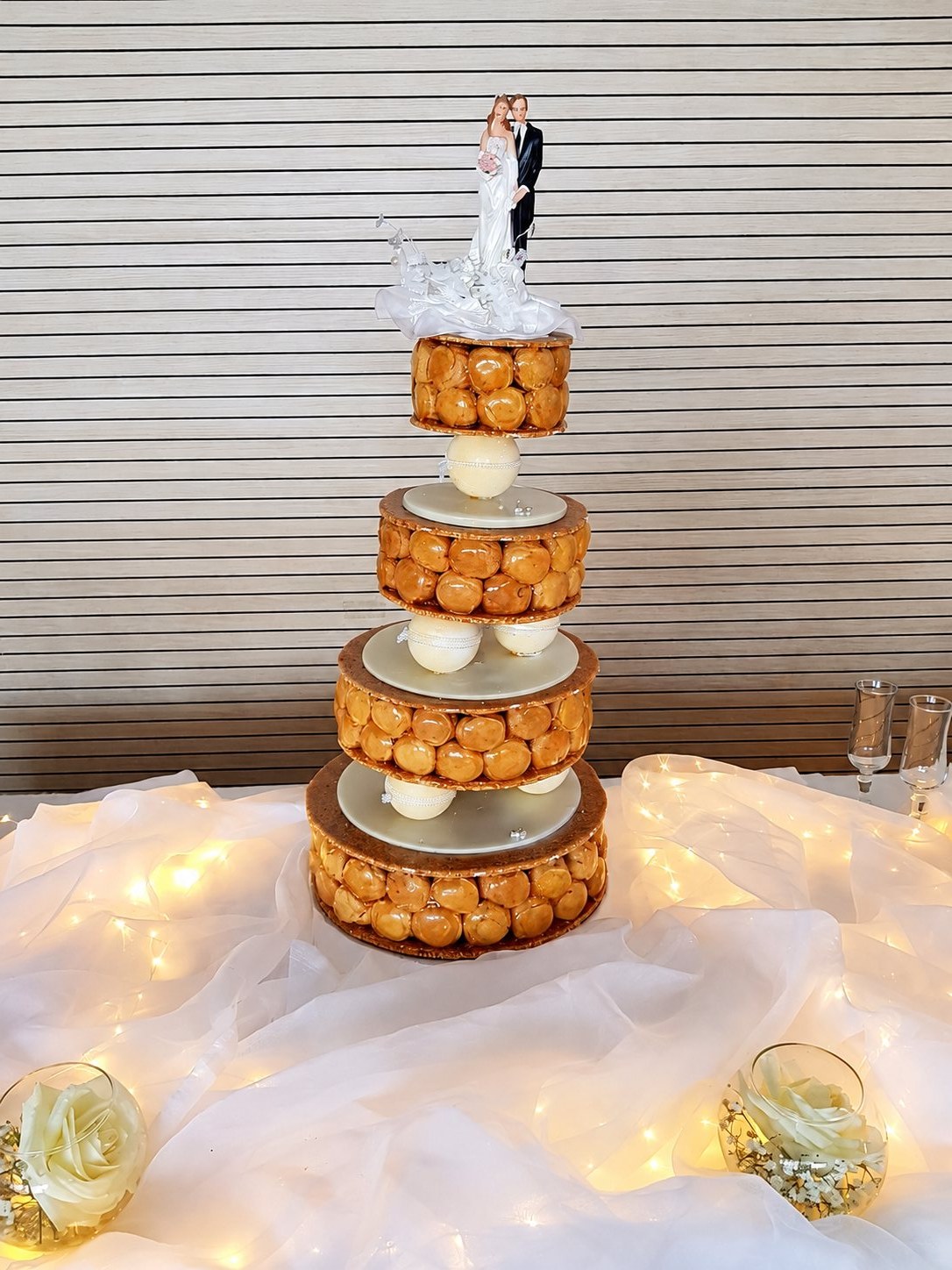 Une pièce montée à quatre étages de choux caramélisés, surmontée de figurines de mariés, est posée sur une table ornée de voiles blancs, de guirlandes lumineuses et de roses.