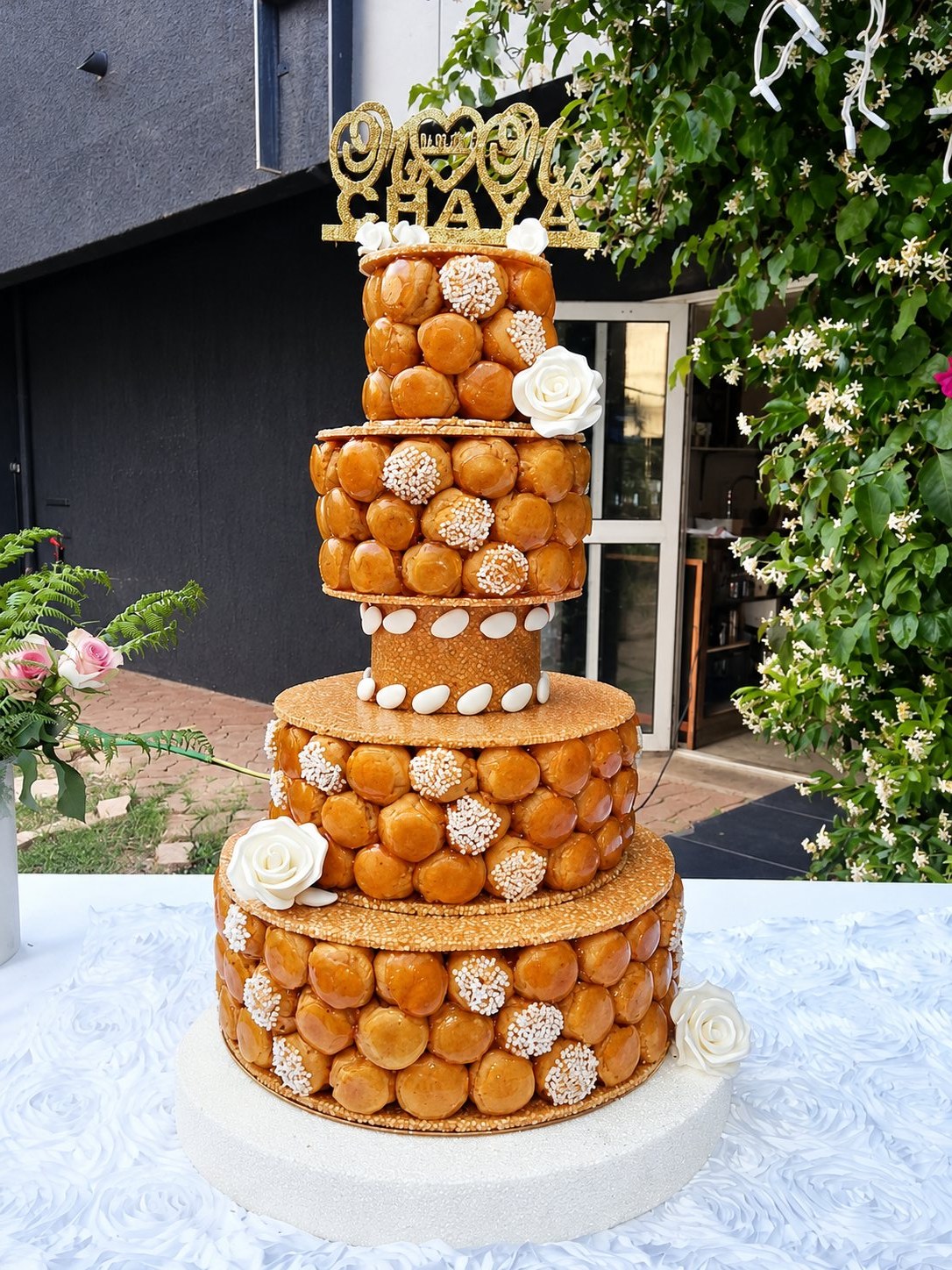 Une pièce montée à trois étages de choux caramélisés ornés de sucre perlé et de roses blanches, posée sur une table dehors devant un jasmin en fleurs.