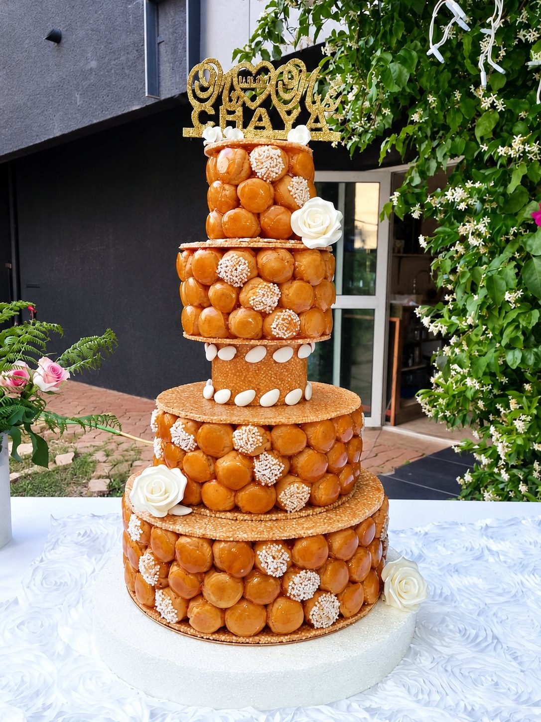 Une pièce montée à plusieurs étages de choux caramélisés, ornée de roses blanches et de dragées, posée sur une table avec nappe blanche dans un jardin fleuri.