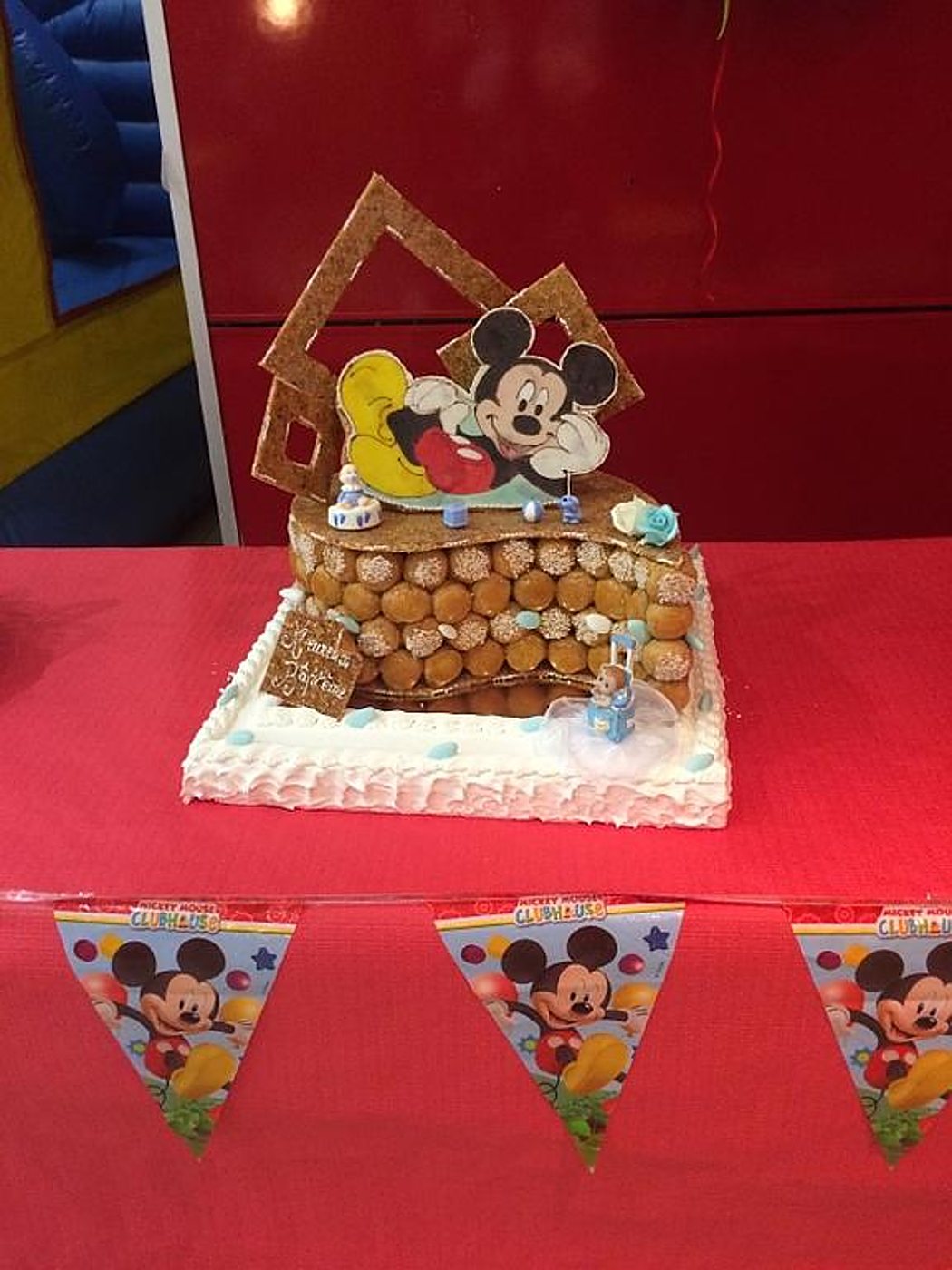 Une pièce montée en choux ornée d'une décoration Mickey Mouse est posée sur une table rouge, devant une guirlande de fanions Mickey Mouse Clubhouse.