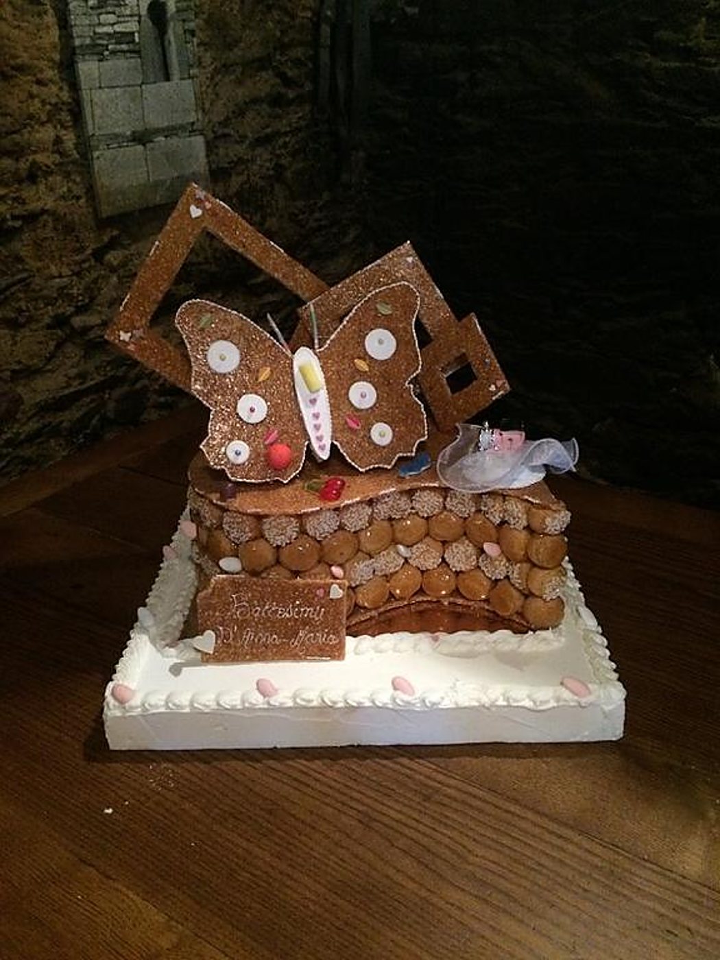 Une pièce montée de choux caramélisés surmontée d'un grand papillon décoré et de cadres en nougatine, posée sur une table en bois devant un mur de pierre.