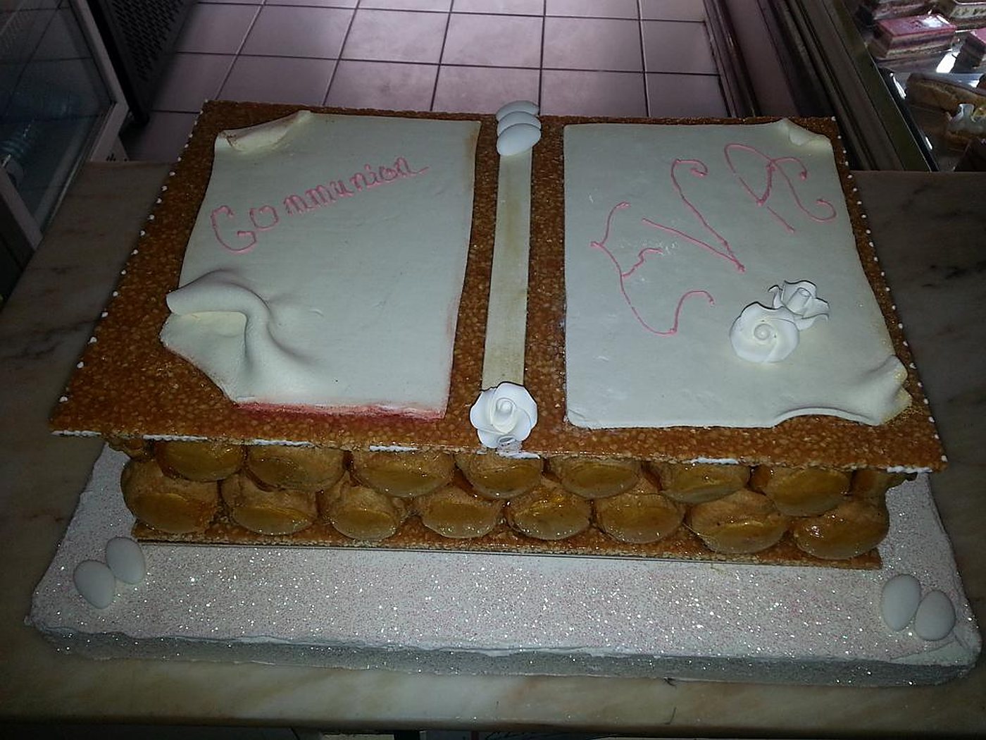 Un grand gâteau de communion en forme de livre ouvert, composé de choux caramélisés, de pages en pâte blanche ornées de roses en sucre et de dragées, posé sur un comptoir en marbre.