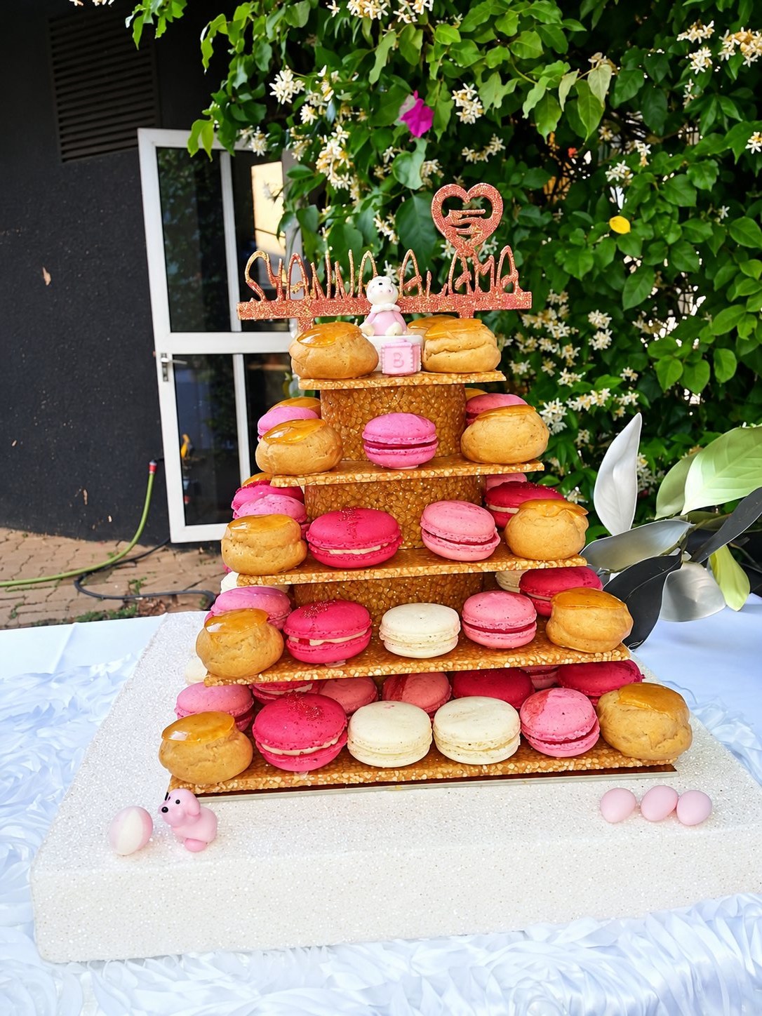Une pièce montée en gradins garnie de macarons roses et blancs et de choux caramélisés, posée sur une table dans un jardin fleuri.