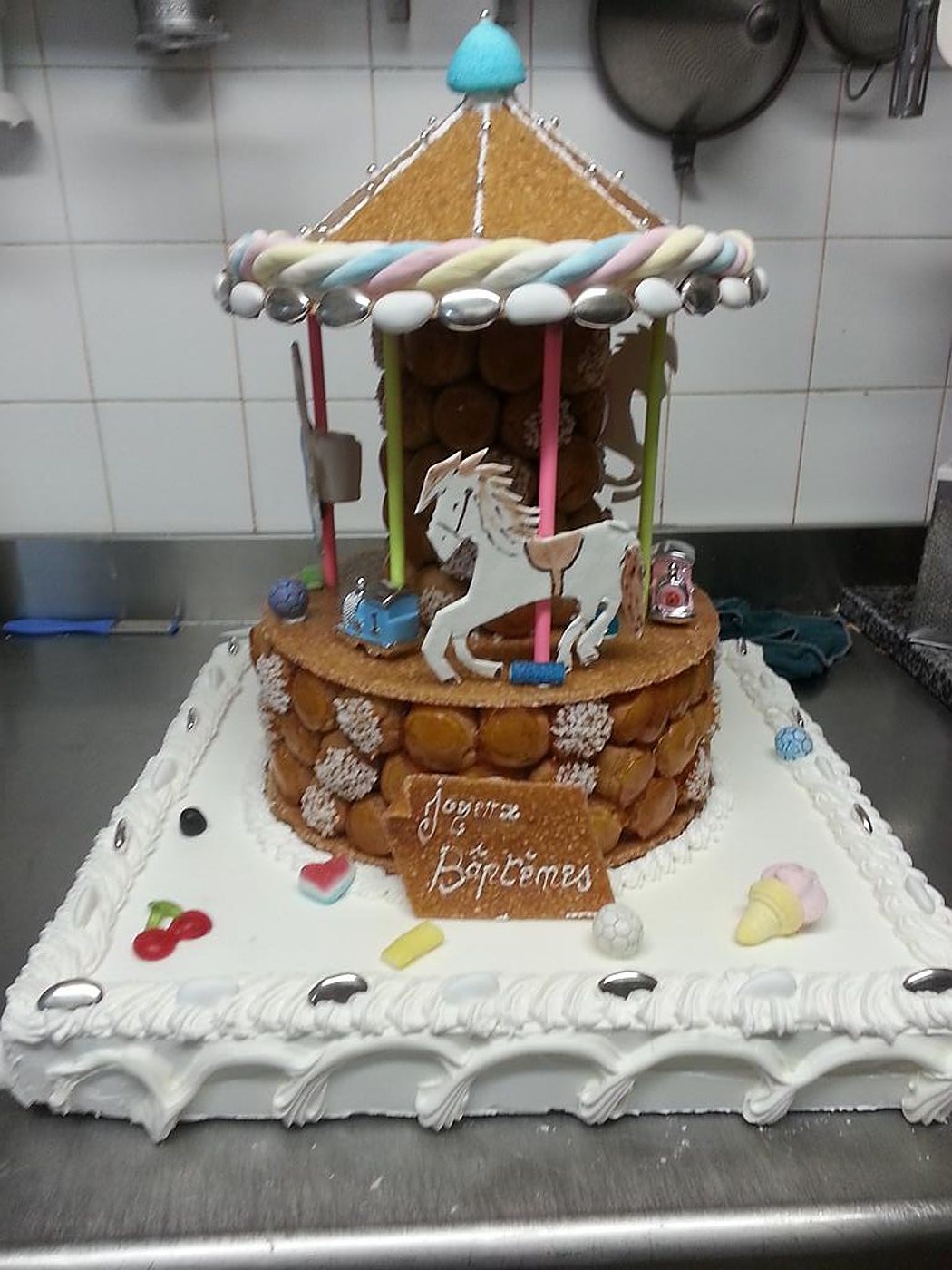 Une pièce montée en forme de manège de choux caramélisés, décorée de bonbons, de guimauves torsadées et d'un cheval en sucre, posée sur un plateau de crème blanche pour un baptême.