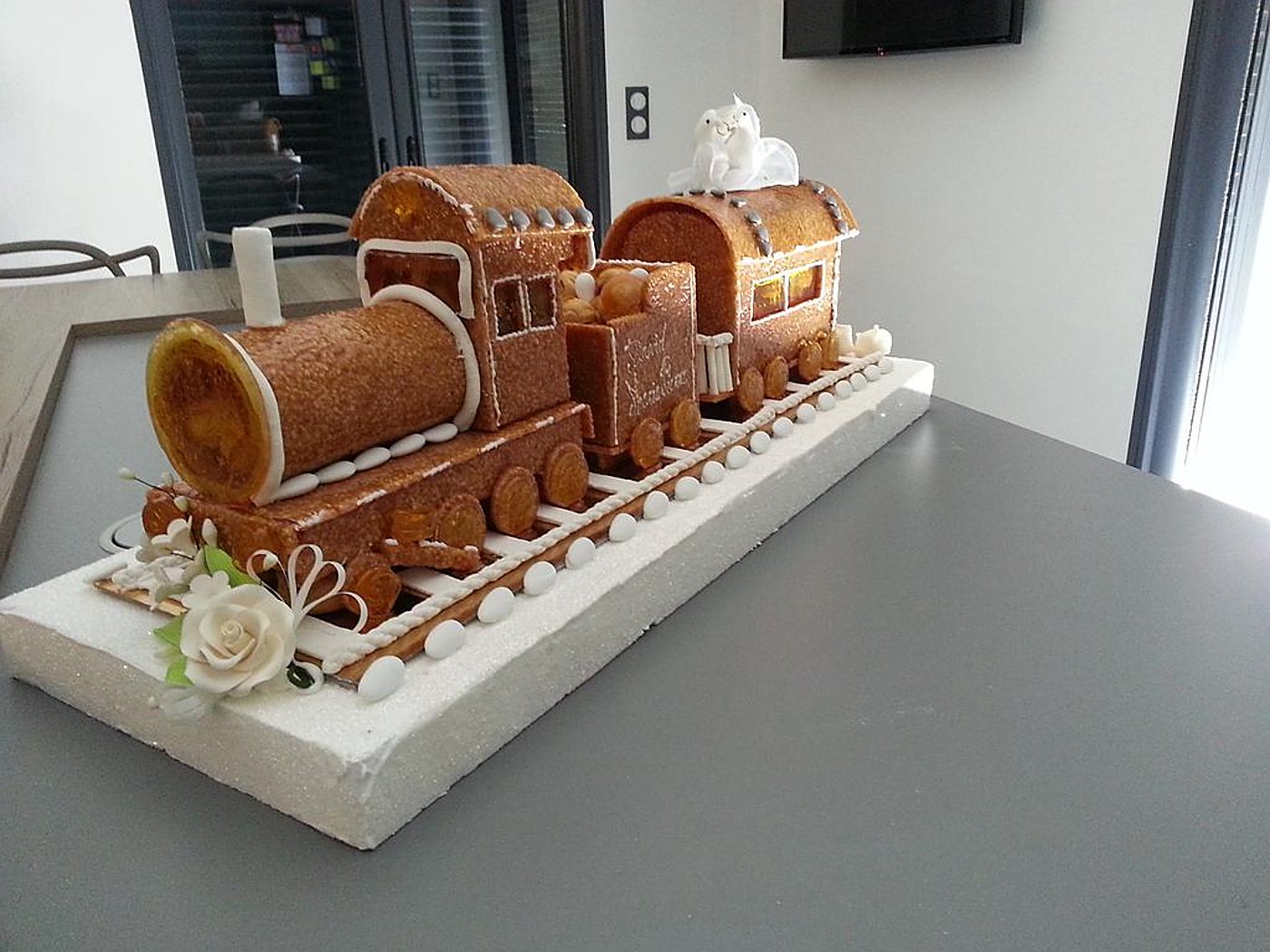 Une pièce montée en forme de locomotive et de wagons sur des rails en sucre, ornée d'une rose blanche et de deux colombes, posée sur une table grise dans une pièce.
