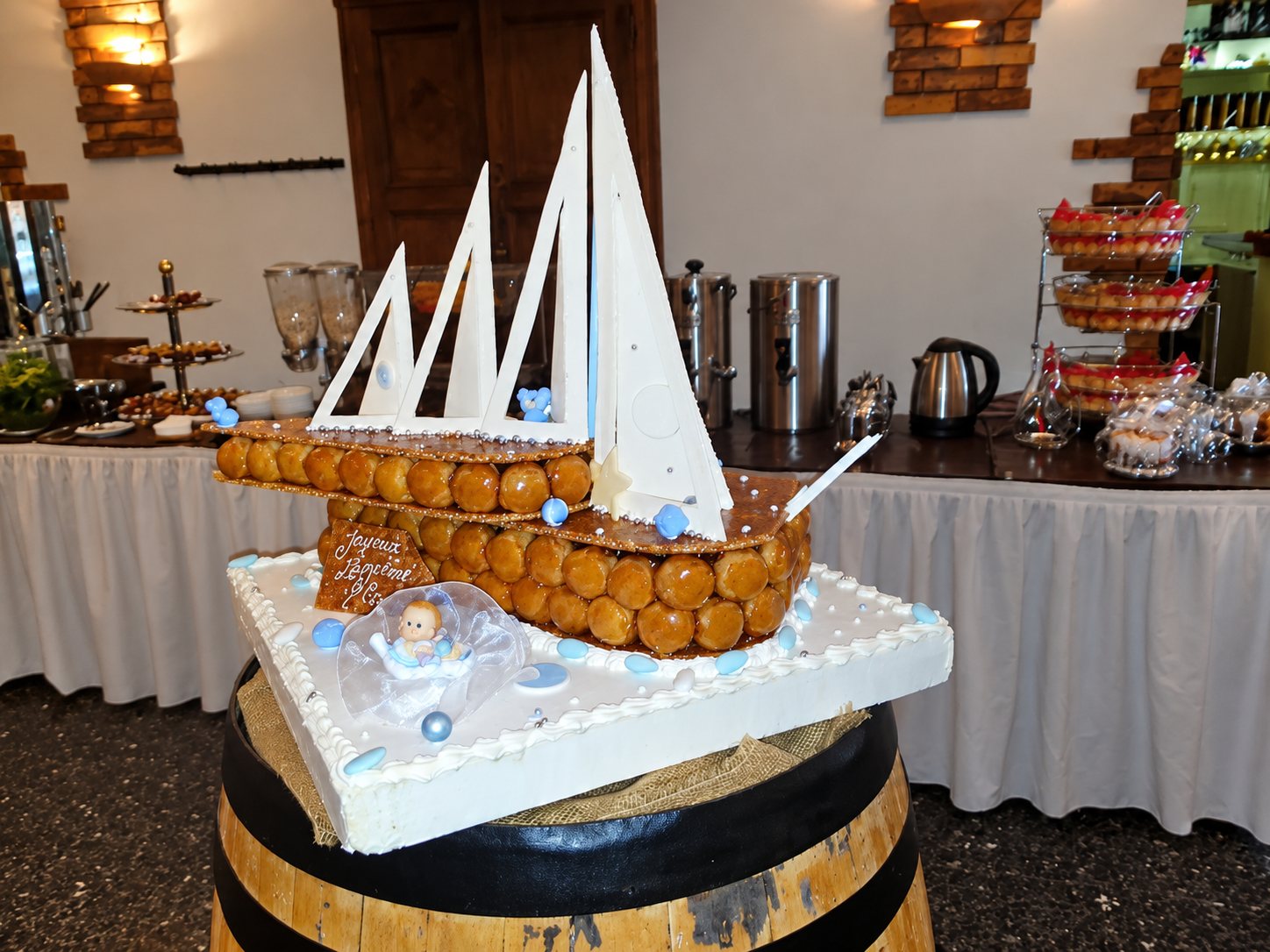 Une pièce montée en forme de voilier, faite de choux caramélisés avec des voiles blanches, est posée sur un tonneau devant un buffet de réception.