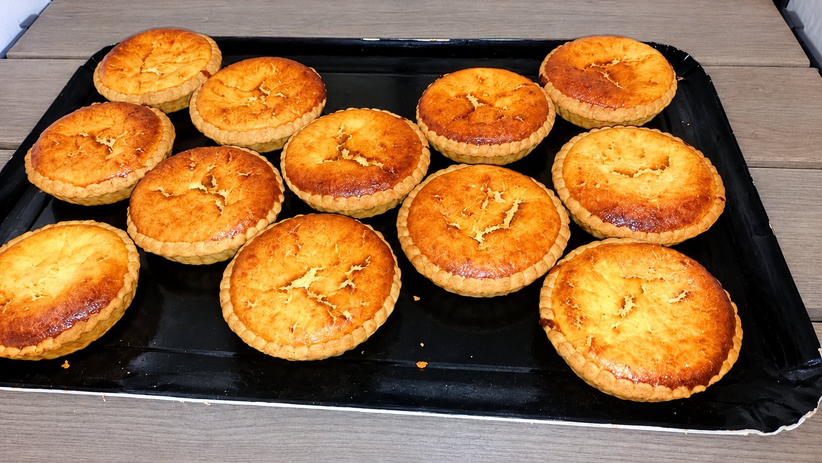 Douze petites tartelettes à la surface dorée et craquelée, posées sur un plateau en carton noir placé sur une table en bois gris.