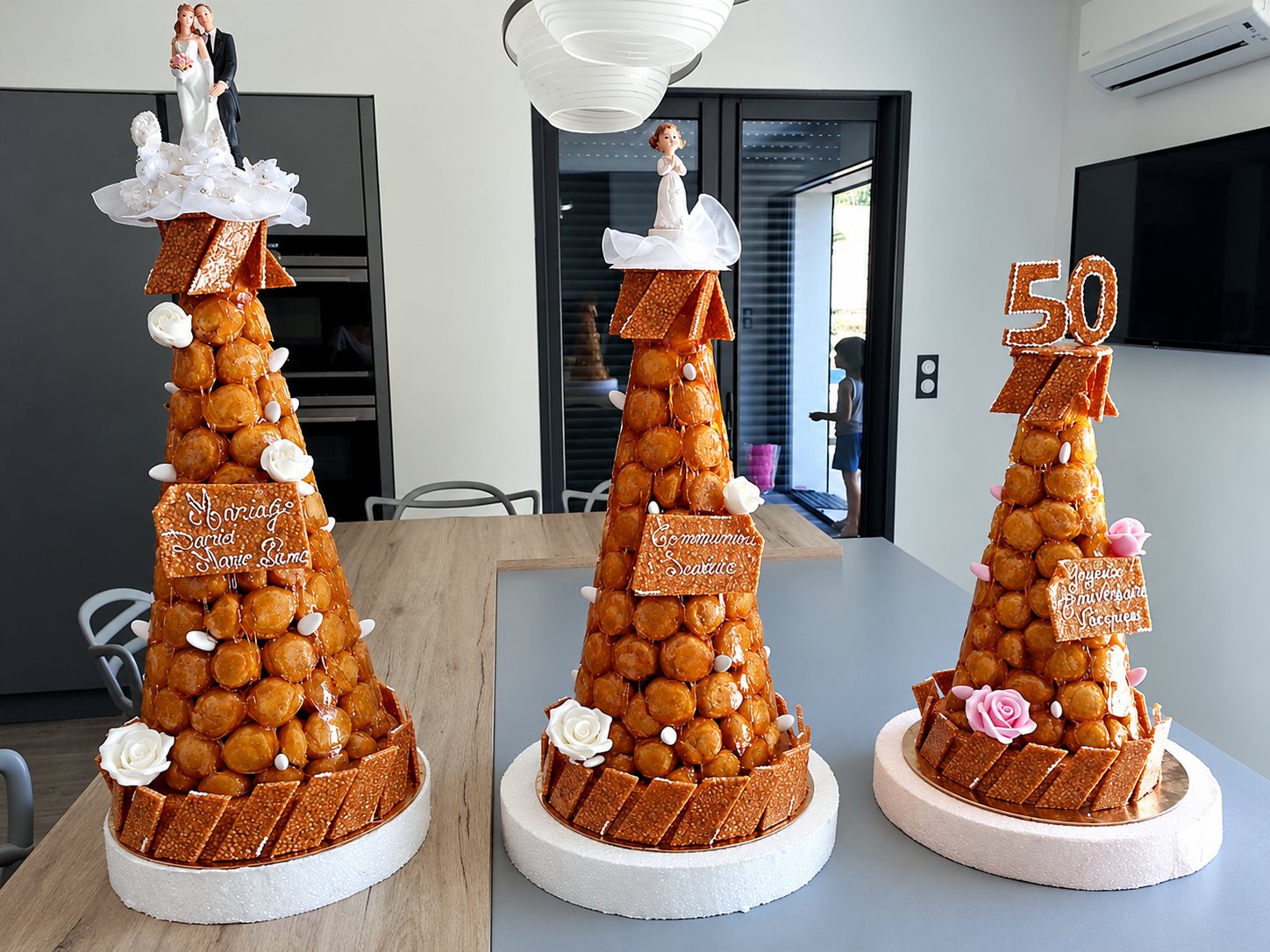Trois pièces montées en choux caramélisés, décorées de roses en sucre et de figurines, posées sur une table dans une pièce lumineuse.